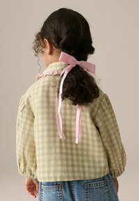 Chemise à carreaux vert et crème avec des manches bouffantes et des accents en ruban rose au niveau du col, associée à un jean en denim bleu.