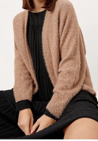 Beige huňatý cardigan s otevřeným předním dílem, texturovanou tkaninou a černým lemem na manžetách, nošený přes černé plisované šaty.