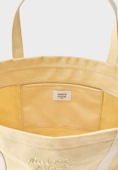 Sac fourre-tout en toile beige avec poche à fermeture éclair ouverte à l'intérieur et étiquette "Maison Kitsuné Paris" cousue sur la poche.