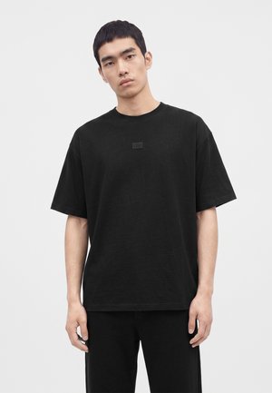 Jeune homme aux cheveux courts noirs portant un t-shirt noir uni oversized et un pantalon noir sur un fond clair uni.