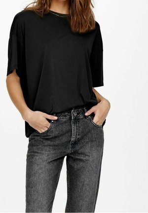 Kvinde iført en løs sort T-shirt stoppet ned i højtaljede, slidte sorte jeans med hænderne i lommerne på en hvid baggrund.