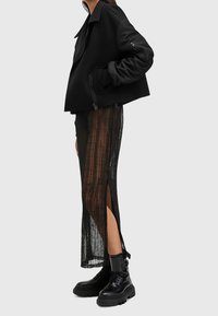Femme portant une veste courte noire, une jupe en maille noire transparente avec une fente latérale, et des bottes noires épaisses à lacets, posée de profil sur un fond neutre.