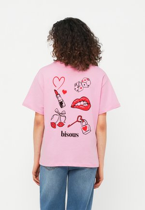 Kvinne med krøllete hår iført en rosa T-skjorte med røde lepper, leppestift, terninger, hjerter, en nøkkel med et hjerteformet lås, og ordet "bisous".