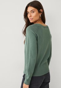 Maglione verde in un materiale morbido con scollatura rotonda, maniche lunghe, orlo a coste e silhouette aderente. Nessun motivo o accento visibile.