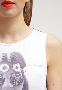 Bílé tričko bez rukávů s tištěnou černobílou grafikou ženy s květinovými slunečními brýlemi a květinovým věncem. Měkký bavlněný materiál.