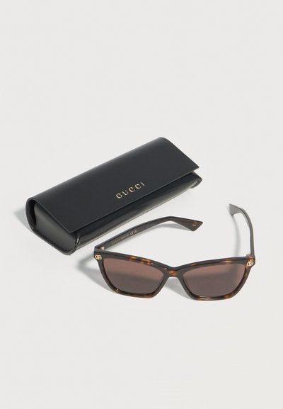 Ορθογώνια γυαλιά ηλίου σε χρώμα χελώνας με σκουρόχρωμους φακούς και χρυσό λογότυπο Gucci, δίπλα σε μαύρη δερμάτινη θήκη Gucci με χρυσή σήμανση.