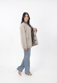 Cárdigan de punto beige con un ajuste relajado, que presenta grandes botones negros, dos bolsillos frontales, puños de canalé y una textura suave.