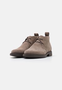 Botas de tobillo de ante marrón con punteras redondeadas, costuras mínimas, cintas delgadas y un pequeño lazo en la parte trasera para facilitar su uso. Suela de goma plana.