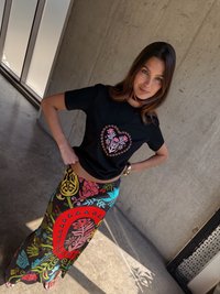 Μαύρη μπλούζα με σχέδιο λουλουδιού σε σχήμα καρδιάς σε ροζ και κόκκινο, συνδυασμένη με μια μακριά, πολύχρωμη φούστα με γεωμετρικά σχήματα και έντονα λουλουδάτα σχέδια.