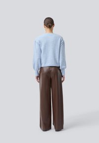 Helle blaue, weiche Strickjacke mit ballonförmigen Ärmeln, kombiniert mit hoch taillierten, weiten braunen Lederhosen. Der Hintergrund ist neutral grau.