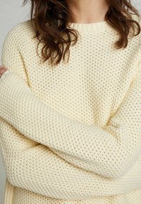 Kermanvalkoinen neulepaita, jossa on teksturoitu, avoin kudoskuvio sekä resorit hihansuuissa ja pääntiessä. Kangas näyttää pehmeältä ja mukavalta.