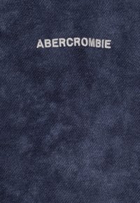 Tela azul marino con una textura suave que presenta la palabra "ABERCROMBIE" bordada en hilo blanco. Diseño simple y directo.