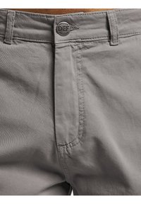 Pantalon gris avec devant plat, fermeture par bouton métallique avec logo "DEF", poches cousues et tissu lisse avec une texture subtile.
