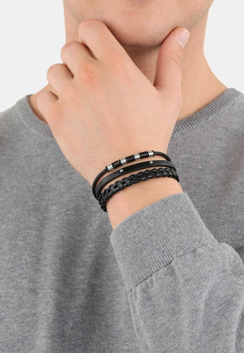 Pulseras de cuero negro con texturas variadas; una cuenta con detalles metálicos. Llevada en una muñeca, combinada con un suéter gris.