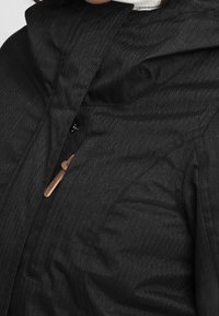 Detalní záběr na černý kabát v herringbone vzoru s límcem a koženým zapínáním na přední straně.