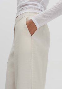 Beige tailored pants with a smooth texture, front pockets, and a slightly relaxed fit. Worn with a light gray long-sleeve shirt.