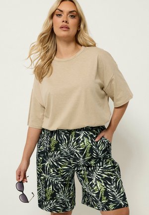 Vrouw met lang blond haar, een beige T-shirt met korte mouwen en zwarte korte broek met groene en witte bladpatronen, die een zonnebril vasthoudt.