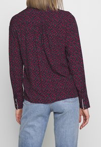 Blusa a maniche lunghe in tessuto blu navy scuro con un motivo floreale sparso in rosso e bianco, dotata di polsini con bottoni e una vestibilità morbida.