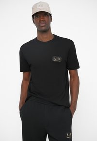 T-shirt preta com mangas curtas e um emblema ao peito com um logótipo dourado. Combinada com calças de treino pretas. Um boné bege completa o conjunto.