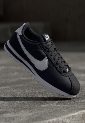 Sneaker en cuir noir avec des accents gris, bout arrondi, surface texturée et semelle extérieure blanche. Caractéristiques : laçage central et logo sur la languette.