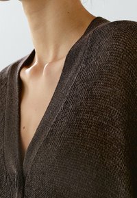 Close-up of a person’s neck and upper chest wearing a dark brown textured knit cardigan with a deep V-neck.