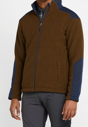 Uomo che indossa una giacca in pile marrone con toppe blu navy sulle spalle e sui gomiti, con cerniera chiusa, abbinata a pantaloni grigio scuro.