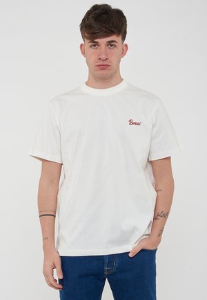T-shirt corto a maniche corte color crema realizzato in cotone, con un piccolo logo ricamato rosso sulla parte sinistra del petto. Il modello indossa jeans blu.