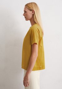 Senfgelb Bluse mit kurzen Ärmeln aus leichtem Stoff, mit rundem Ausschnitt, lockerer Passform und kleinen seitlichen Schlitzen am Saum.