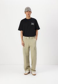 Camiseta negra de algodón con texto impreso, pantalones cargo beige, gorra gris y zapatillas blancas con suelas marrones. Estilo casual, corte relajado.
