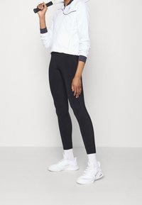 Biała kurtka z ciemnoniebieskimi akcentami, czarne legginsy i białe sneakersy. Kurtka ma zamek z przodu i prążkowane mankiety, natomiast legginsy są dopasowane.