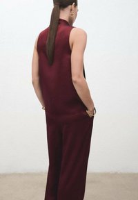 Mono sin mangas en burdeos con un cuello alto, textura suave, bolsillos laterales y un corte holgado, acompañado de accesorios en tono dorado.