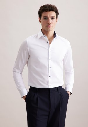 Weißes Hemd mit navyfarbigem Akzentkragen, langen Ärmeln und dunklen Knöpfen; kombiniert mit dunklen Hosen, glatte Stofftextur.
