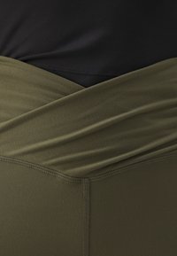 Leggings vert olive avec une large ceinture pliée portée sur un haut noir ajusté, montrant la texture du tissu et les détails des coutures.