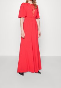 Maxi dress in bright red fabric with short puff sleeves and lace detailing at the neckline, featuring a fitted waist and flowing bottom.