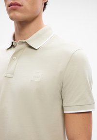 Polo couleur kaki en coton, avec un col classique, des accents blancs et un logo carré avec "BOSS" sur la poitrine. Manches courtes.