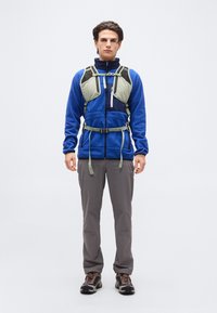 Άνδρας φοράει μπλε φλις μπουφάν, γκρι παντελόνι πεζοπορίας, καφέ παπούτσια πεζοπορίας και ανοιχτό πράσινο θώρακα με ιμάντες και αγκράφες.