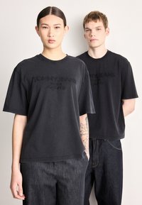 Tricouri negre din bumbac cu mâneci scurte, având logo-ul „TOMMY JEANS” în relief. Modelele poartă blugi din denim închis la culoare, evidențiind o croială casual.