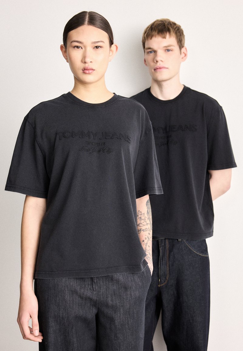 Tricouri negre din bumbac cu mâneci scurte, având logo-ul „TOMMY JEANS” în relief. Modelele poartă blugi din denim închis la culoare, evidențiind o croială casual.