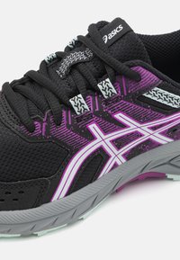 Zapatilla deportiva negra con detalles en púrpura y blanco, que cuenta con una parte superior de malla texturizada, lengüeta acolchada y suela de caucho gris con patrón de tracción.