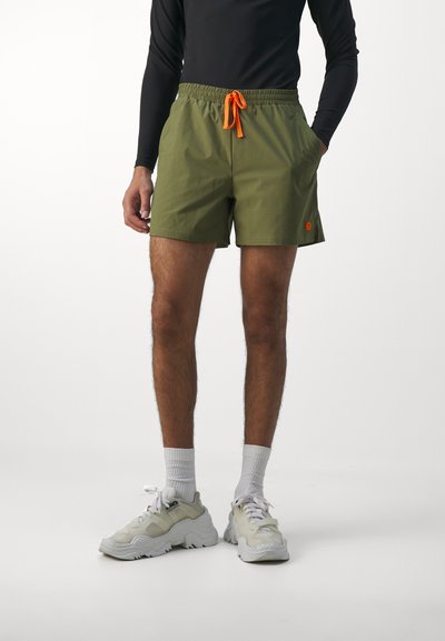 Shorts de bain verts avec une taille élastique et un cordon de serrage orange. Portés avec des chaussettes blanches à la cheville et des baskets beiges épaisses.
