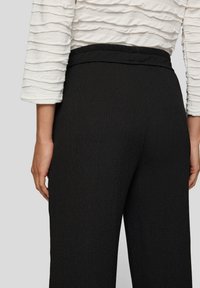 Schwarze, strukturierte Hosen mit weitem Bein, ausgestattet mit einem taillierten Bund und dezenten Bügelfalten, kombiniert mit einem weißen, crêpierten Langarmoberteil.