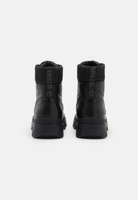 Botas negras hasta el tobillo con un exterior de cuero brillante, cuello de tela texturizada y suela gruesa con detalles de logo en relieve.