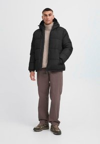 Giacca a vento nera con cappuccio, texture a trapunte, cerniera frontale e tasche laterali, abbinata a un dolcevita beige e pantaloni marroni.