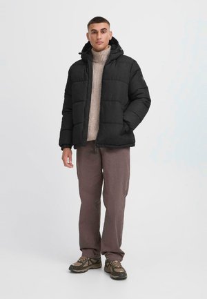 Schwarze Steppjacke mit Kapuze, gequiltete Textur, Frontreißverschluss und seitliche Taschen, kombiniert mit einem beigen Rollkragenpullover und braunen Hosen.