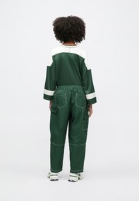 Osoba s kučeravými vlasmi oblečená v tmavozelenej a bielej dlhoruká blúzka, voľné zelené nohavice s vreckami a biele tenisky, stojaca chrbtom.