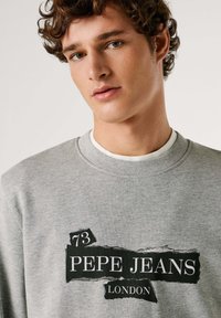 Felpa grigia con collo rotondo, caratterizzata da un logo nero con la scritta "PEPE JEANS LONDON" e un numero "73" in un design a effetto carta strappata.