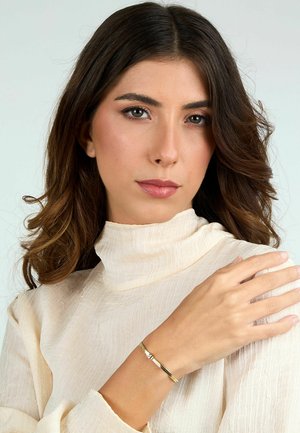 Bracciale color oro con superficie testurizzata e accenti lisci, indossato su un polso, su uno sfondo neutro. Top di tessuto color crema stropicciato.