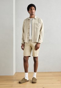 Chemise en lin beige à boutons, t-shirt blanc, short beige, sabots en suède marron et chaussettes blanches. Design simple et coupe décontractée.