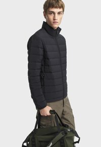 Jeune homme portant une veste matelassée noire et un pantalon beige, tenant un sac de sport vert olive avec des bretelles noires.