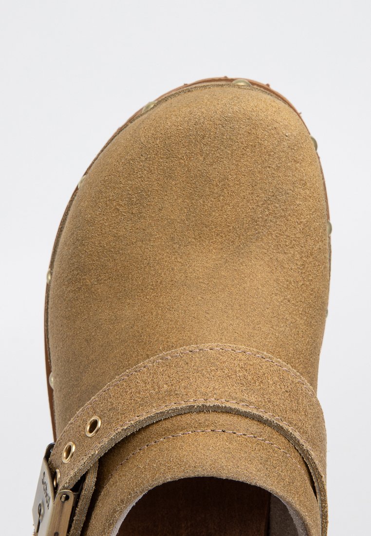 Sabot en suède brun avec un bout arrondi, une boucle ajustable et des accents cousus. La semelle présente de petits points de traction.